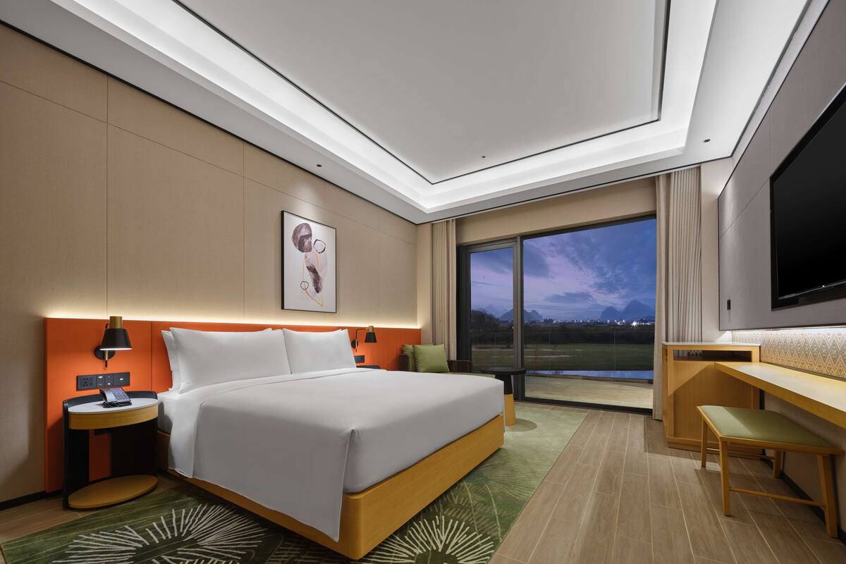 A modern hotel room with a large bed, a desk, a TV, and a view of the outdoors.