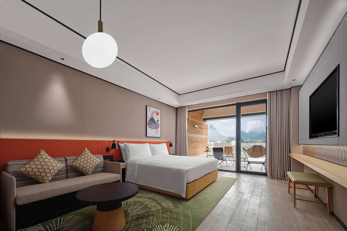 A modern hotel room with a bed, sofa, and a desk. There is a large window with a view of mountains.