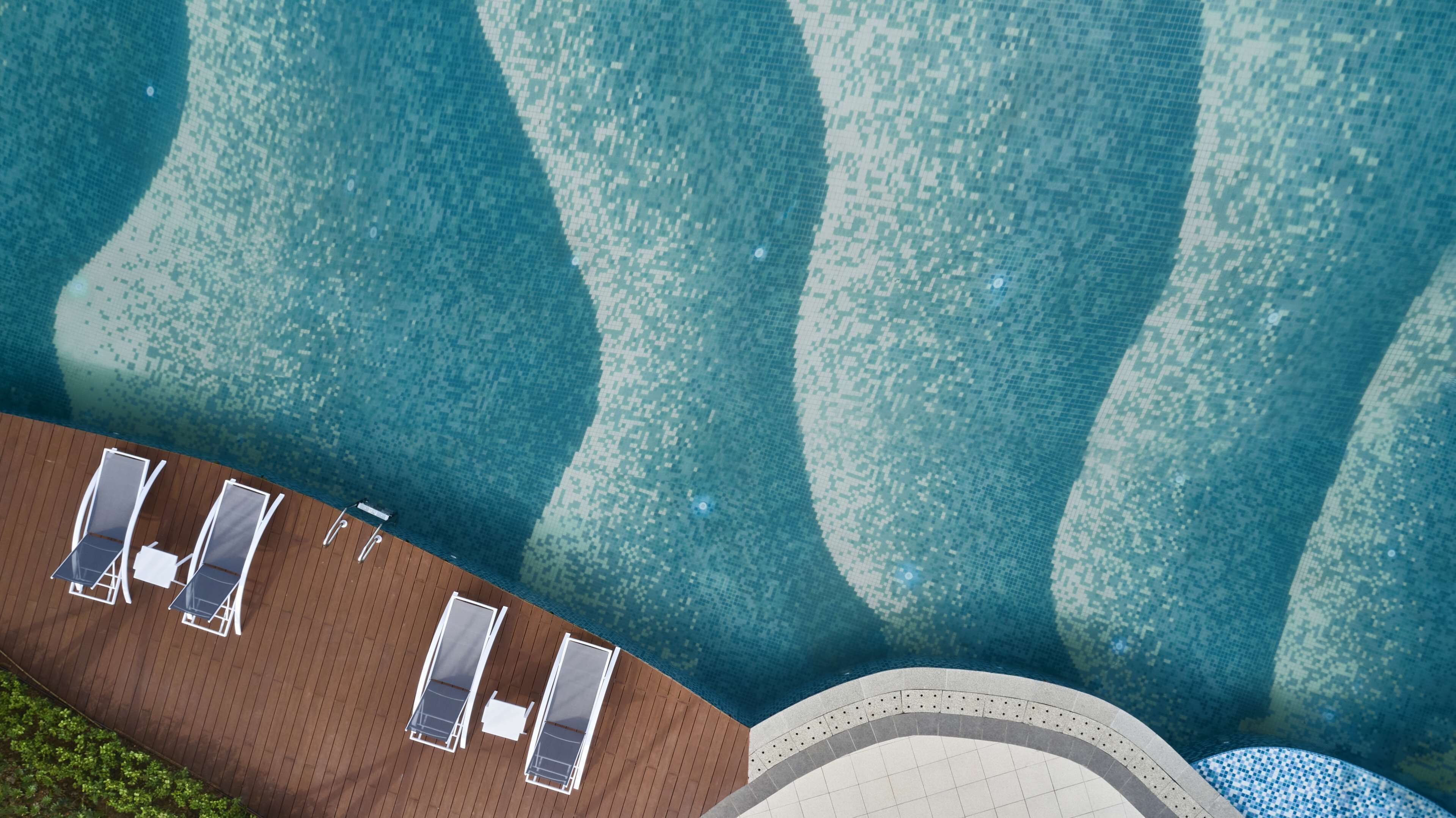 Luftaufnahme eines Schwimmbeckens mit Kachelmustern, einer Holzterrasse und Liegestühlen.