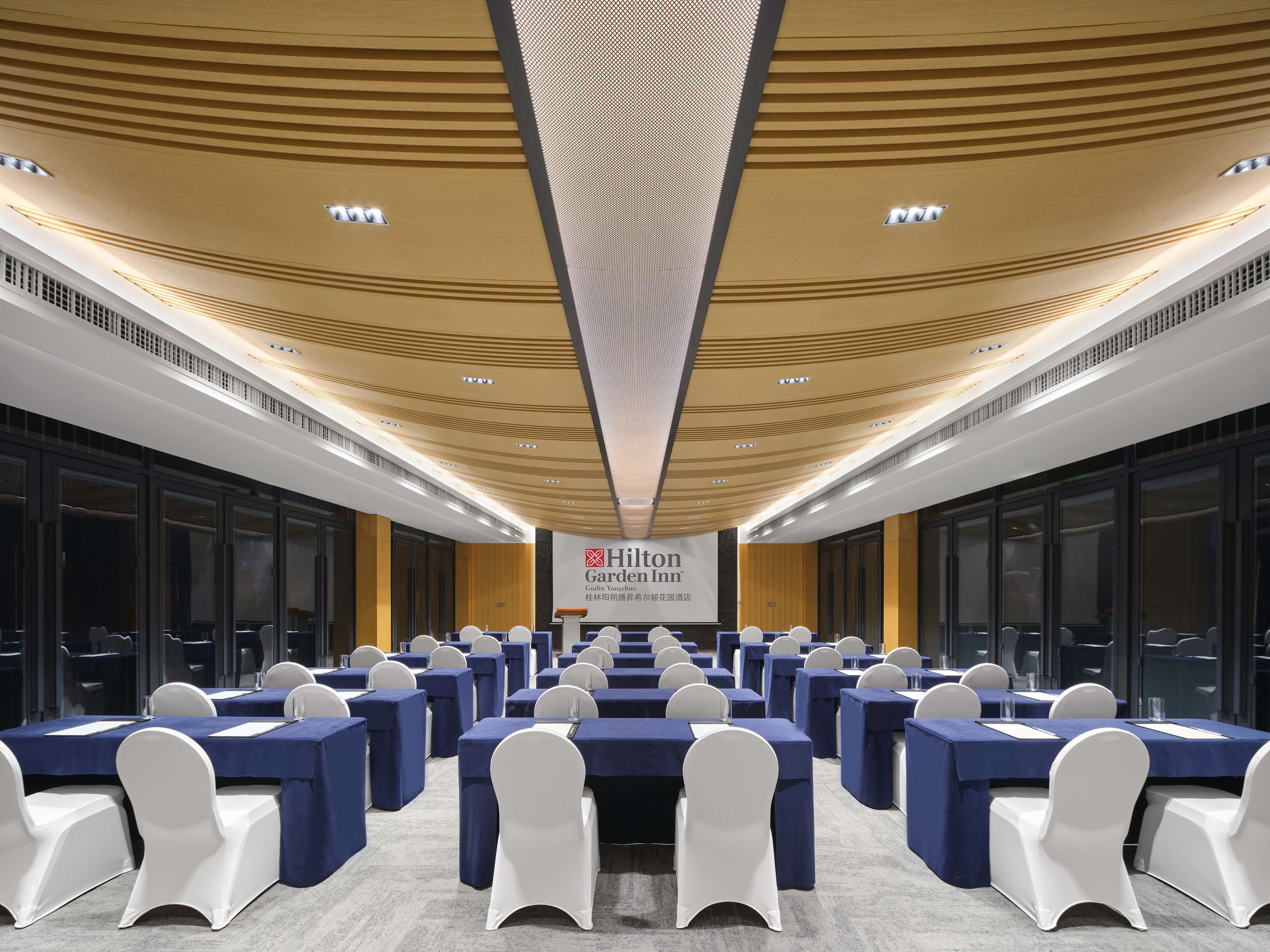 Un salón de conferencias con mesas y sillas, un techo con un diseño único y un cartel de Hilton Garden Inn.