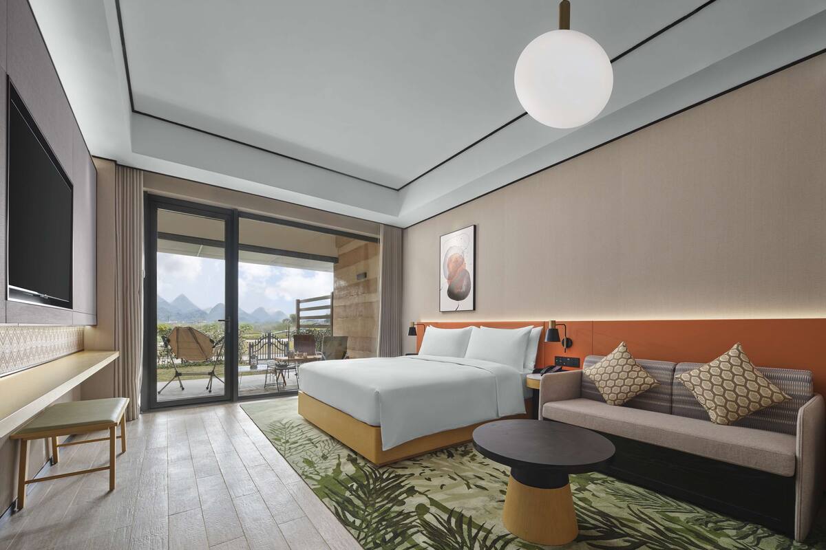 A modern hotel room with a bed, a sofa, a desk, and a large window offering a mountain view.