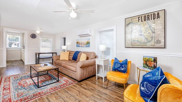 A spacious living room with a wooden floor, a brown sofa, a yellow armchair, and a coffee table. There is a large Florida map on the wall, and a ceiling fan is visible. - Cheerful Coastal Home Close to Beach (Palm Harbor)