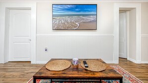 A room with a wooden coffee table, two woven placemats, a remote control, and a framed beach photograph on the wall. - Cheerful Coastal Home Close to Beach (Palm Harbor)