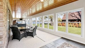 Una habitación amplia con techo de madera y ventanas grandes, una mesa con sillas y vistas a una casa y árboles.