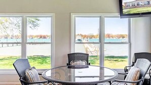 Una habitación con una mesa redonda de vidrio y cuatro sillas de mimbre, ventanas amplias con vista a un cuerpo de agua y un muelle, y un televisor de pantalla plana montado en la pared.