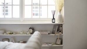 A room with a large window, a shelf with books and decorative items, and a white bed.