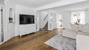 A spacious living room with a large flat-screen TV, a white entertainment unit, a beige sectional sofa, and a staircase leading to another level.
