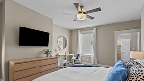 Een slaapkamer met een bed, een bureau, een stoel, een plafondventilator, een televisie en een spiegel.