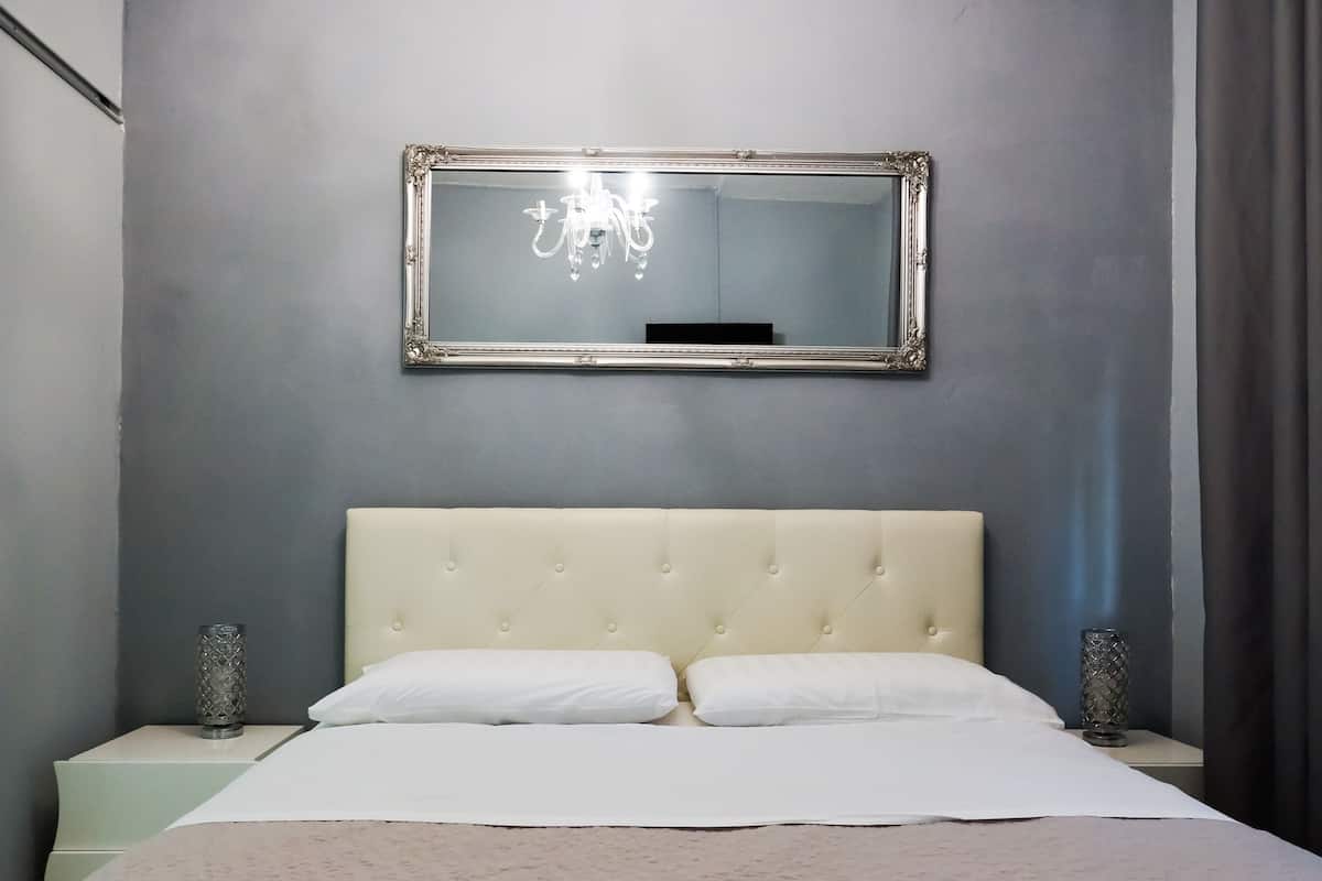 A bedroom with a large mirror, a tufted headboard, two bedside tables with decorative vases, and a bed with white linens.