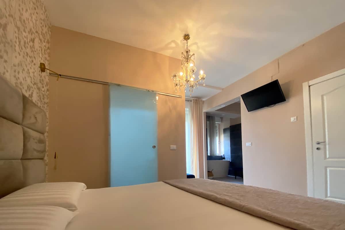A bedroom with a large bed, a chandelier, a flat-screen TV, and a door leading to a bathroom.