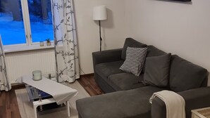 Een woonkamer met een grijze bank, een witte tafel en een raam met gordijnen.