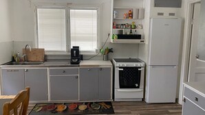 Een kleine keuken met een spoelbak, kookplaat, oven en een koelkast. Er is een raam met zonwering, een aanrecht met diverse keukenbenodigdheden en een houten stoel.
