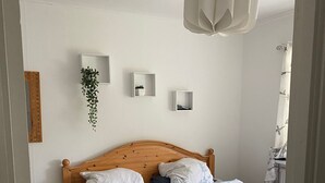 Een houten bed met witte beddengoed in een kamer met witte muren, een hangende plant en een witte hanglamp.