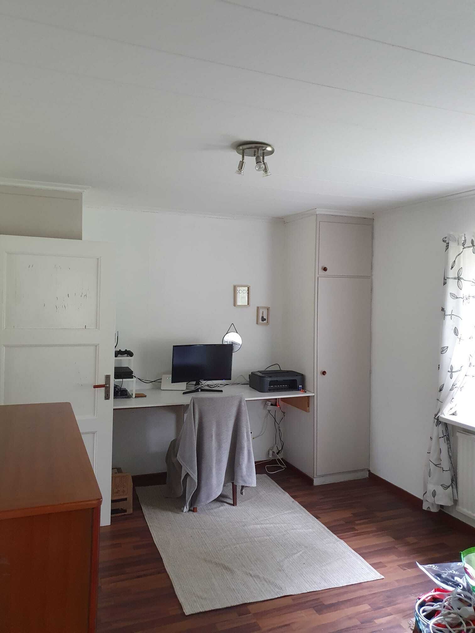 Une petite pièce, peu meublée, avec un bureau, un ordinateur et une imprimante. On y trouve une porte, une petite étagère et une fenêtre avec un rideau.
