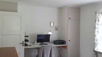 Une petite pièce, peu meublée, avec un bureau, un ordinateur et une imprimante. On y trouve une porte, une petite étagère et une fenêtre avec un rideau.