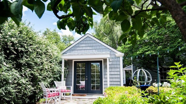 Une petite maison avec une porte d’entrée sombre, des murs extérieurs blancs et un toit en pente. On y trouve un jardin avec différentes plantes et une chaise blanche.