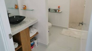 Une salle de bain avec un lavabo noir, des étagères blanches où sont rangés les produits de toilette, des toilettes et un espace douche.