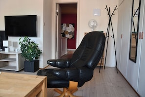 A living room with a black leather chair, a wooden table, a TV, a plant, and a painting on the wall.