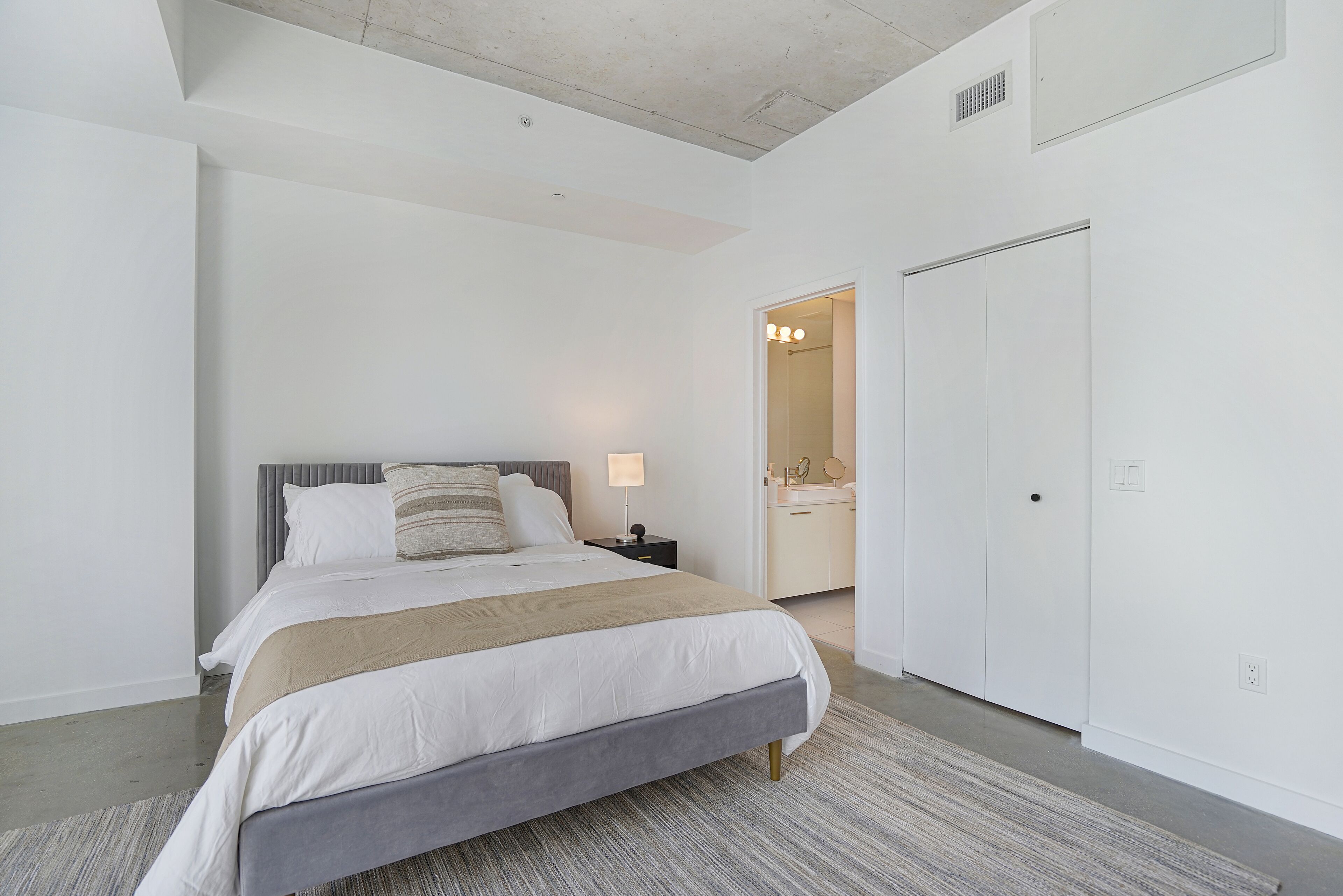Un dormitorio moderno con una cama grande, una mesita de noche, un armario y un baño visible a través de una puerta abierta.