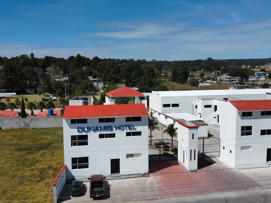 A modern hotel with a red roof, named 'DUNAMIS HOTEL', featuring a parking area and a view of the surrounding landscape.