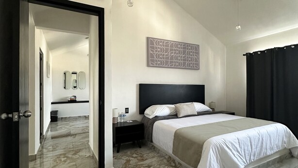 Une chambre d’hôtel moderne avec un grand lit, une table de chevet, une commode, un miroir et une salle de bain visible à travers une porte ouverte.