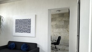 Un salon moderne avec un canapé noir, une table basse en bois et un fauteuil de bureau noir. Un mur blanc est orné d’une œuvre d’art abstraite en noir et blanc, et un mur en pierre se trouve à l’arrière-plan.