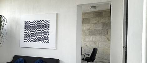 A modern living room with a black sofa, a wooden coffee table, and a black office chair. There is a white wall with a black and white abstract artwork and a stone wall in the background.
