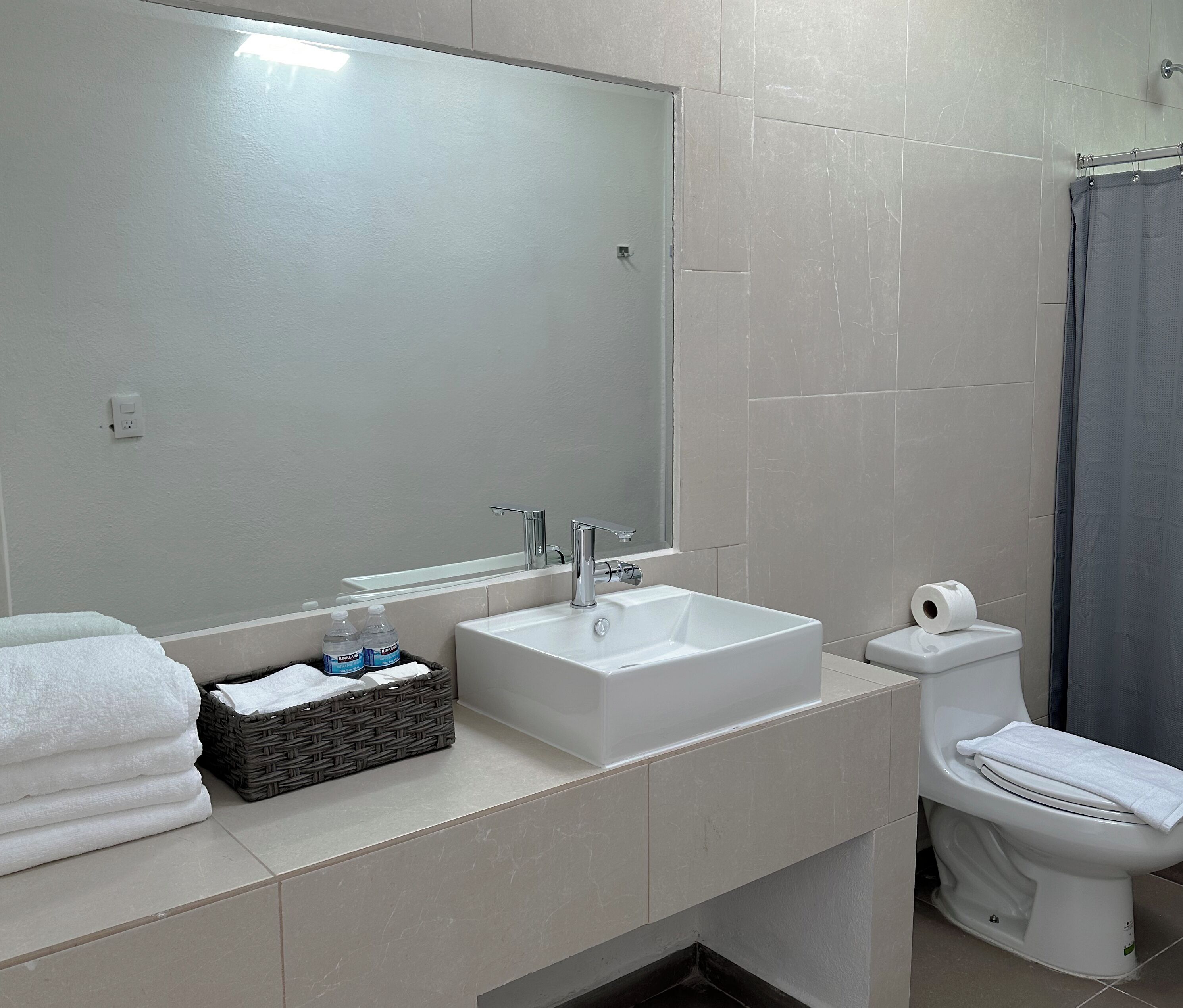 Un baño moderno con lavamanos, espejo y retrete blancos. Hay toallas y artículos de aseo en la encimera.