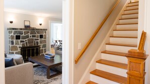 Una escalera con escalones de madera y barandilla, que conduce a una sala de estar con chimenea de piedra, una mesa de centro de madera oscura y vistas al exterior.