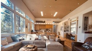Ein großzügiges Wohnzimmer mit einer breiten Eckcouch, einem Hocker und einem Ledersessel. Mehrere Fenster sorgen für natürliches Licht und Blick ins Freie.