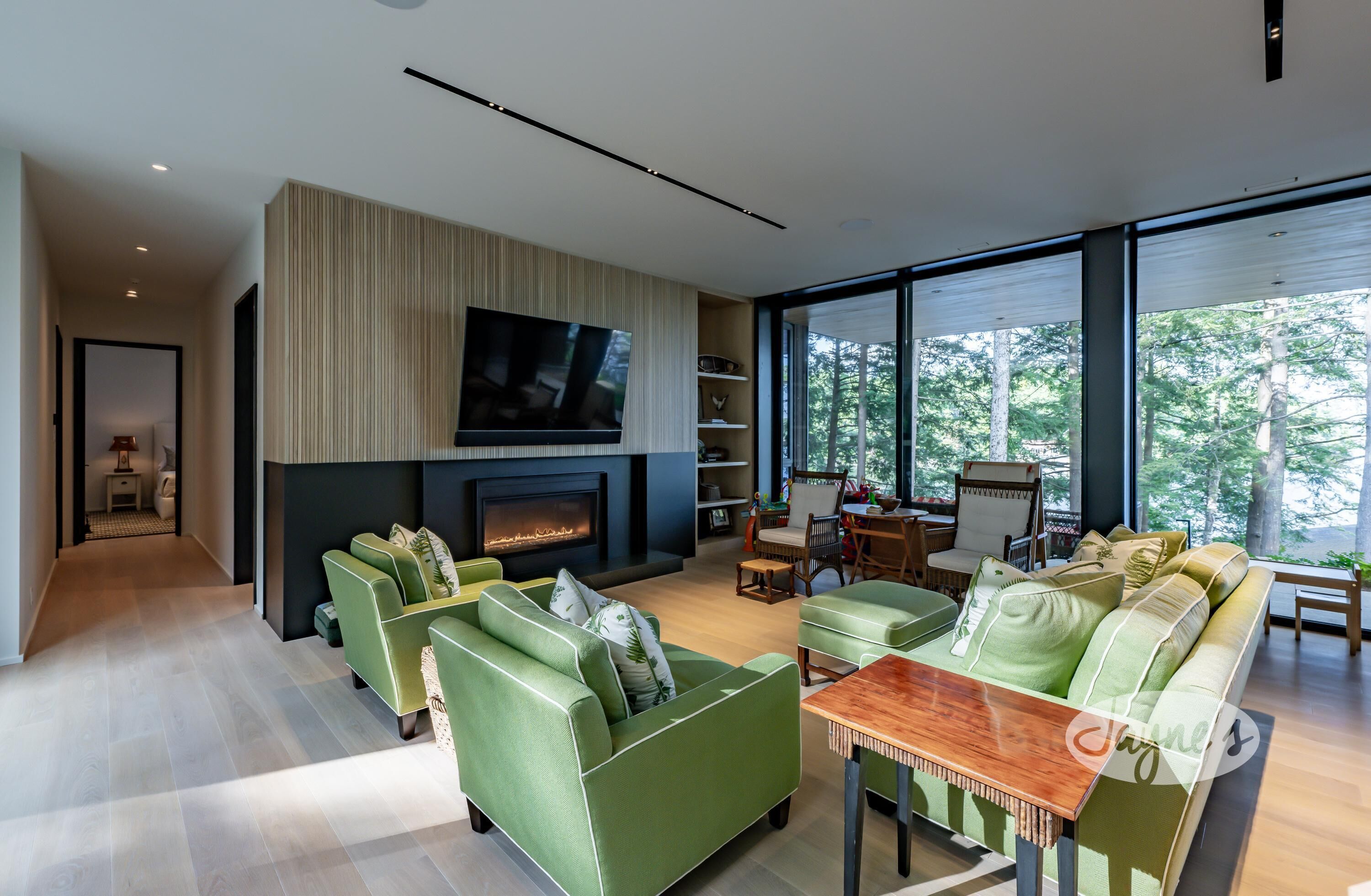 Una sala de estar moderna con sofás verdes, una chimenea, un televisor de pantalla plana y grandes ventanales con vistas a árboles.