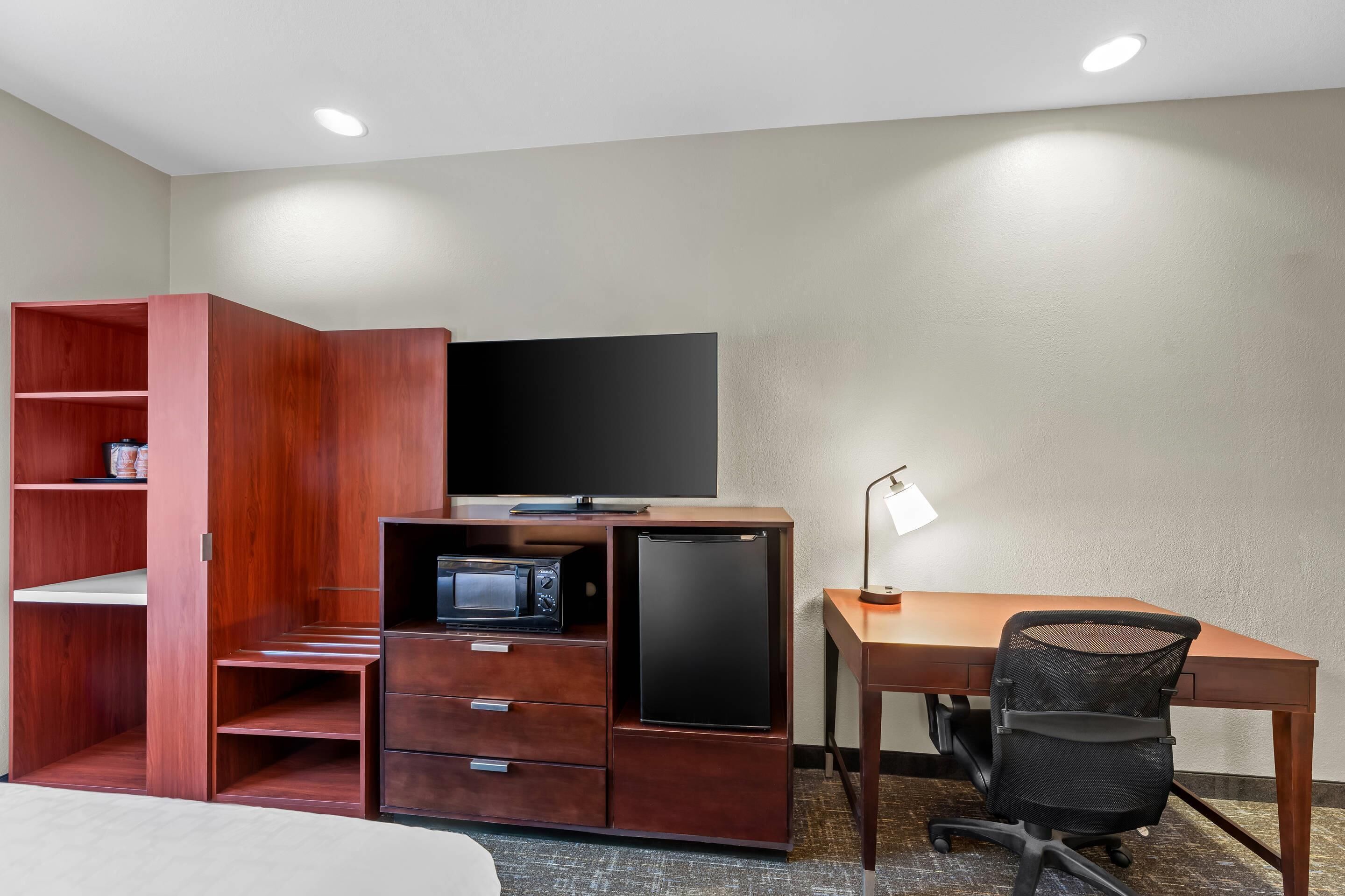 A hotel room with a wooden desk, a television, a microwave, a refrigerator, a chair, and a wardrobe.