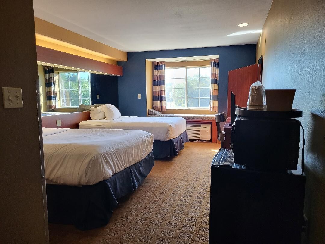 A hotel room with two beds, a desk, a coffee machine, and a window with curtains.