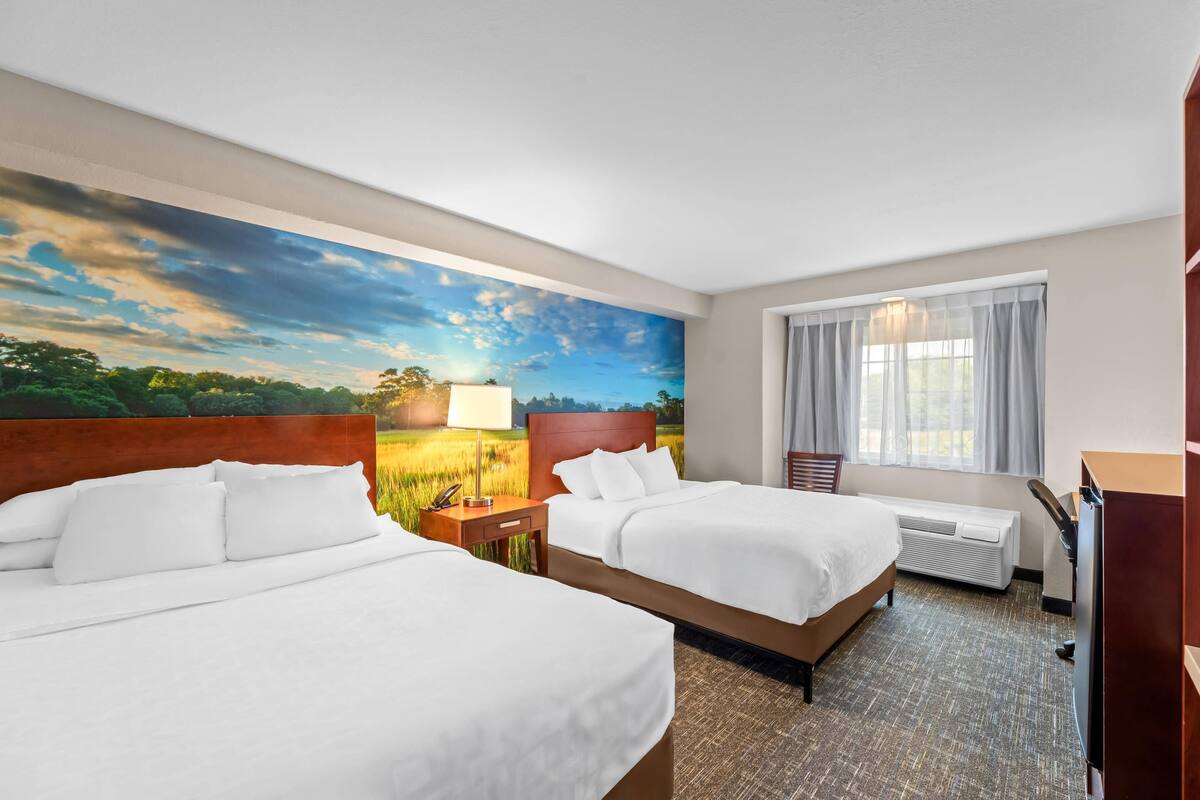 Une chambre d’hôtel avec deux lits, une grande fenêtre avec des rideaux, un tableau représentant un paysage et un bureau avec une lampe.