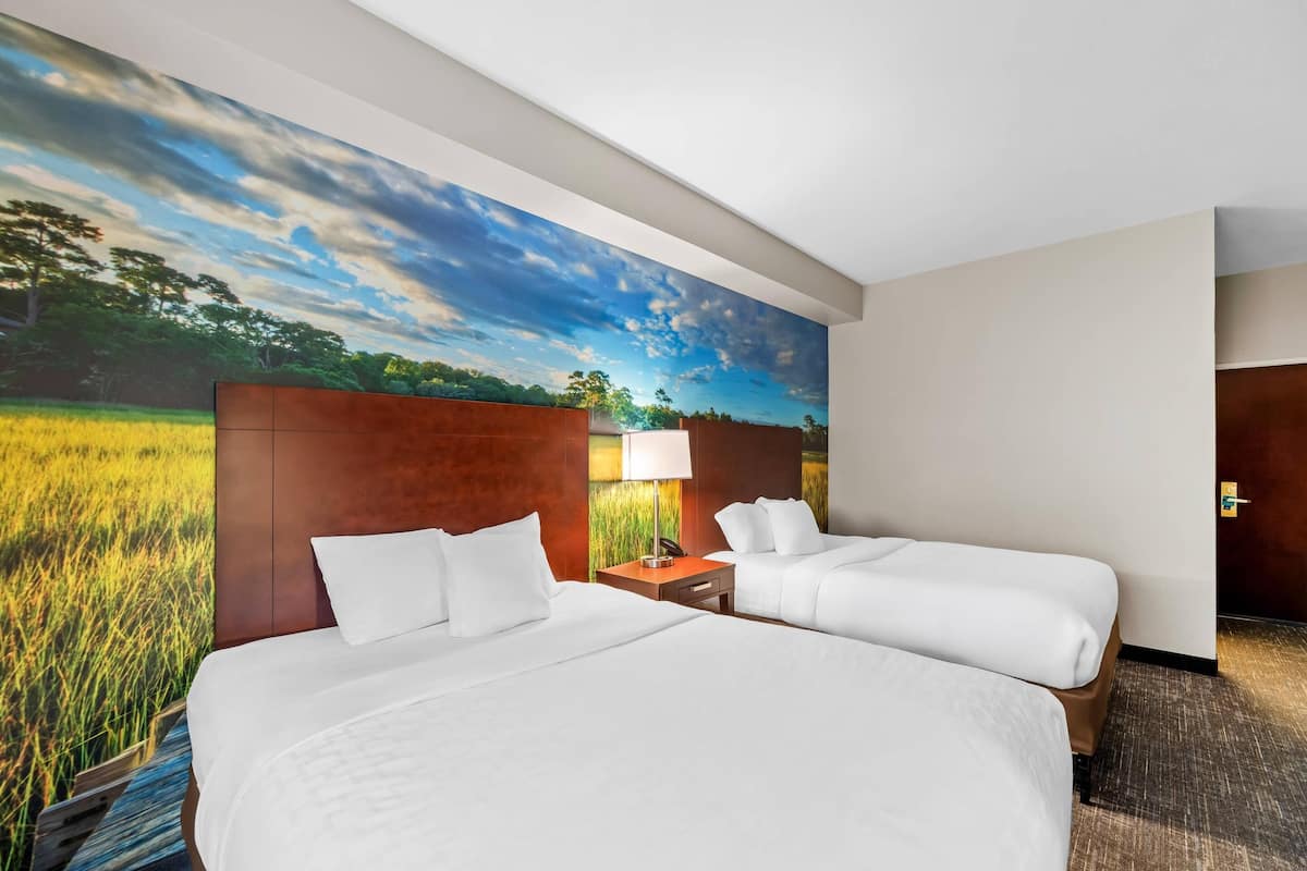 Une chambre d’hôtel avec deux lits, une table de chevet, une lampe et une grande fresque murale représentant un champ et le ciel.