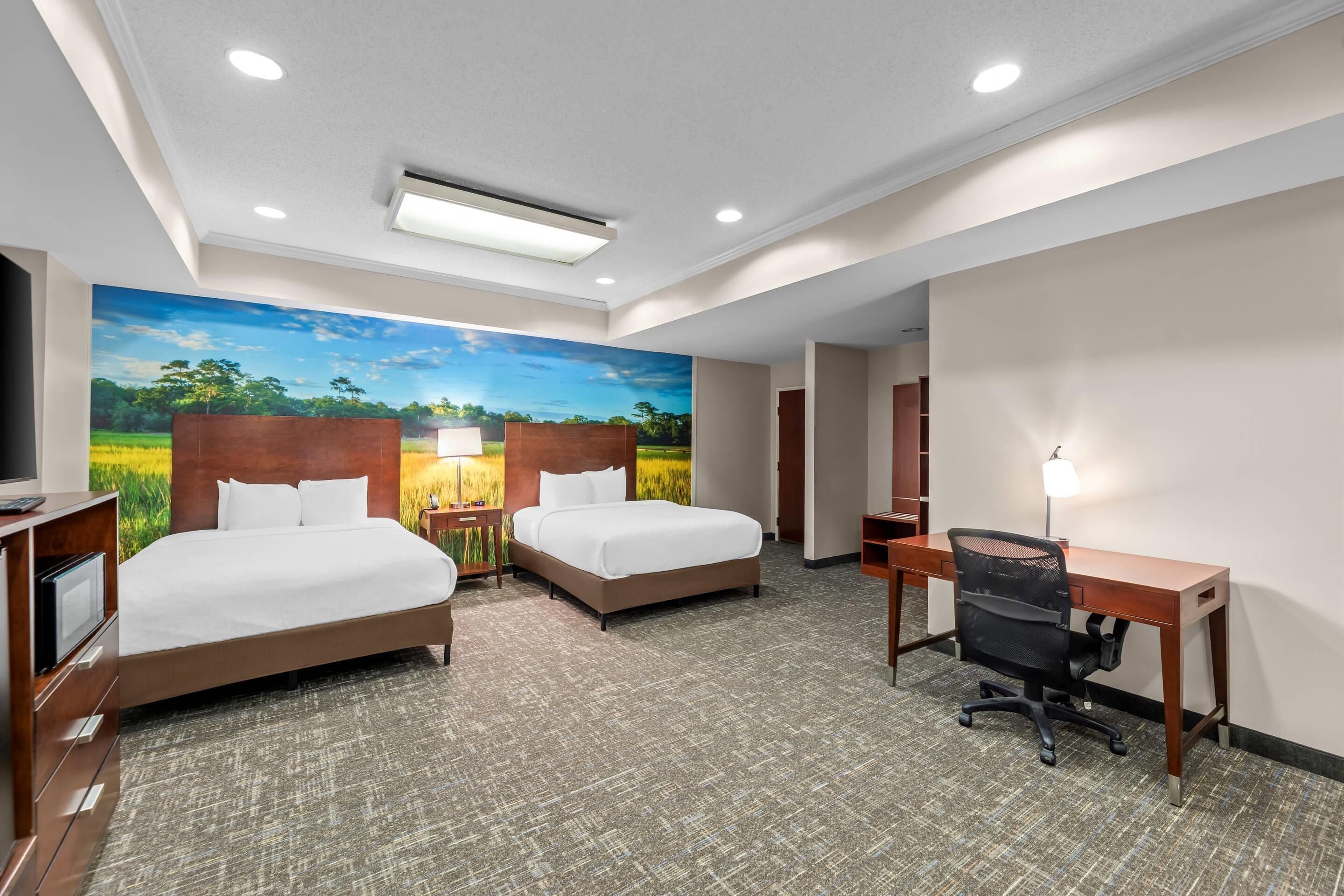 A hotel room with two beds, a desk, a chair, a TV, and a large wall mural.