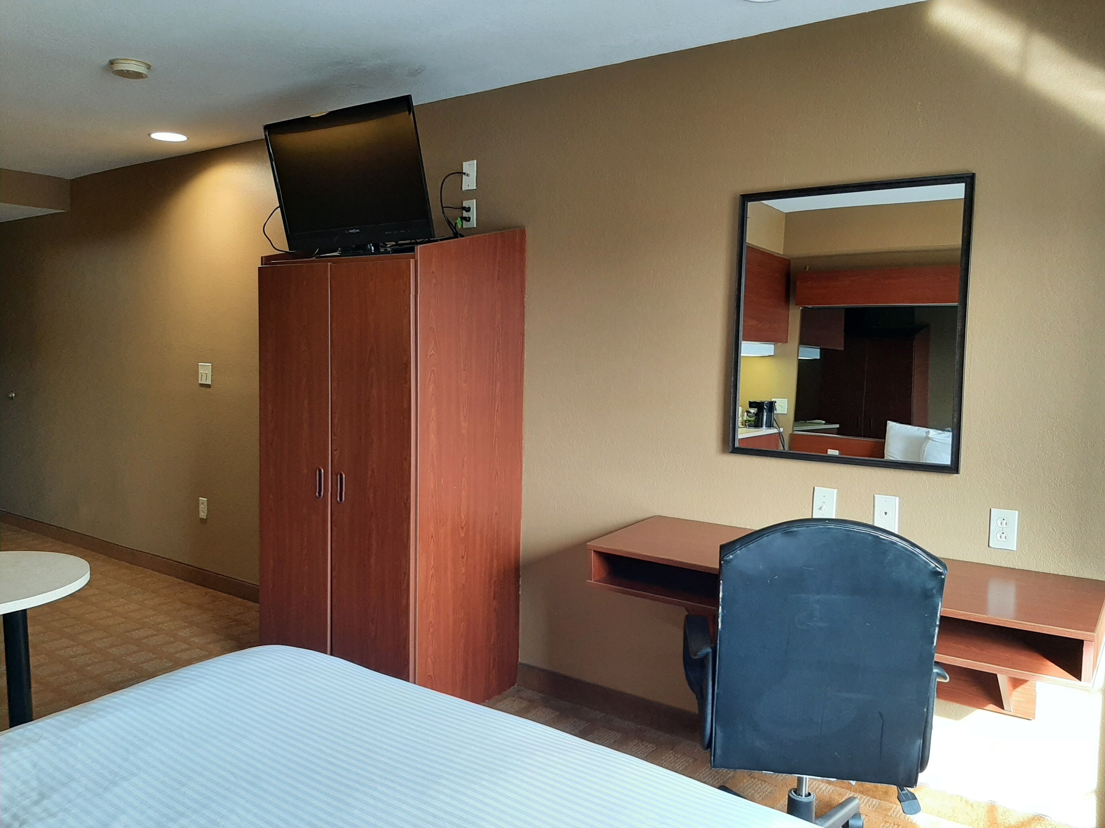 A hotel room with a bed, a desk, a chair, a wardrobe, a mirror, and a television on top of the wardrobe.