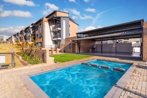 Une piscine avec un patio couvert, une pelouse et un immeuble à plusieurs étages doté de balcons.