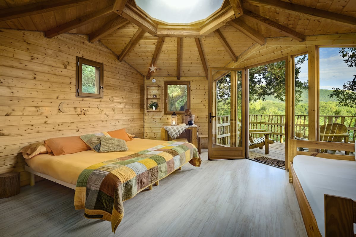 A wooden cabin bedroom with a bed, a nightstand, a window, and a view of the outdoors.