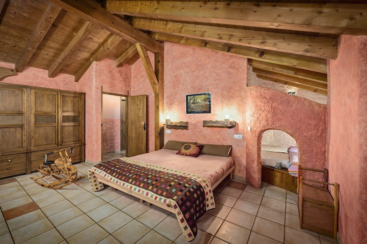A bedroom with a wooden ceiling, a bed with a quilt, a rocking chair, and a wooden wardrobe.