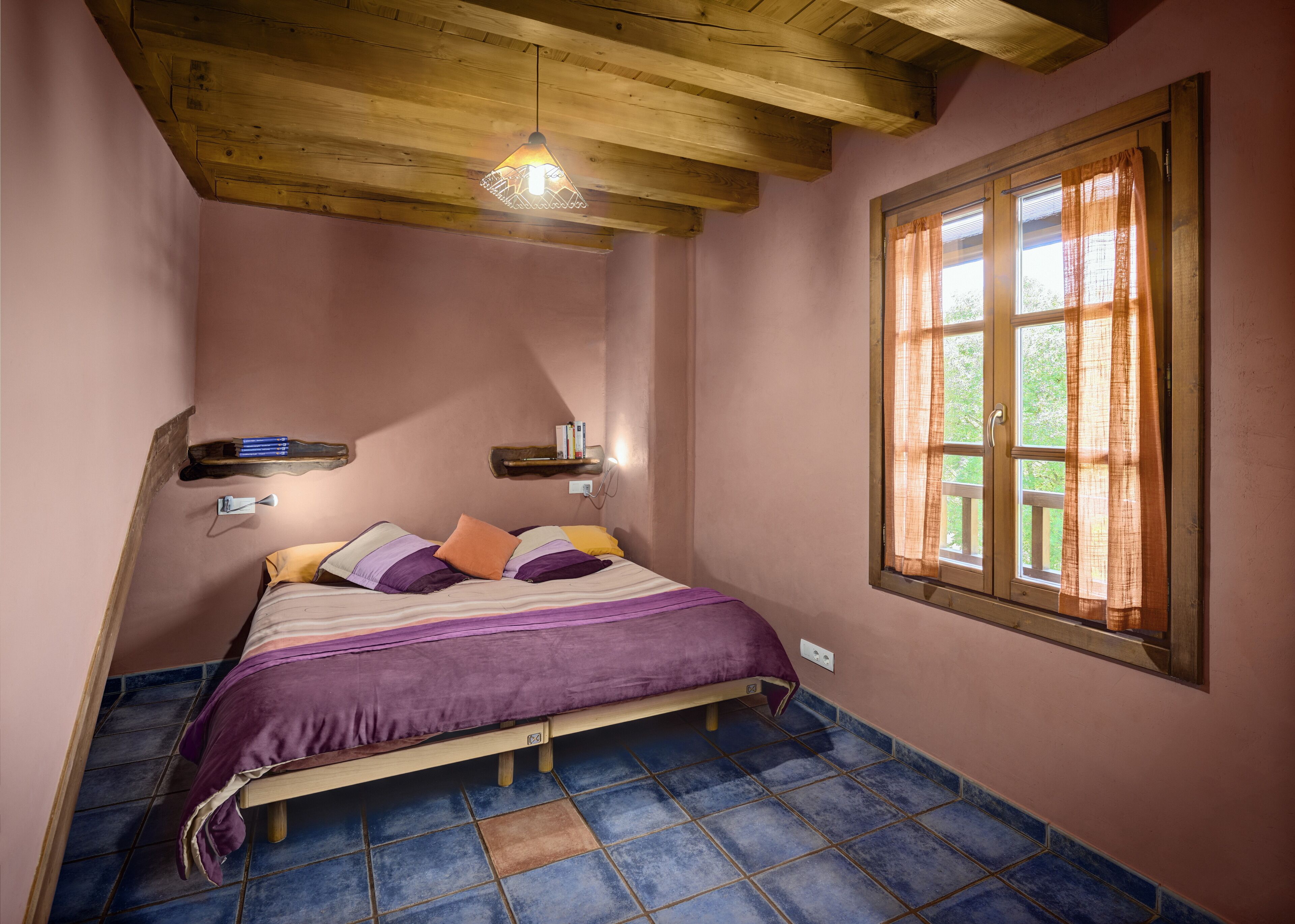 Un dormitorio con techo de madera, una cama con cubrecama púrpura, una ventana con cortinas y un estante pequeño con libros.