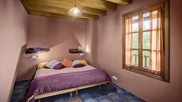 Un dormitorio con techo de madera, una cama con cubrecama púrpura, una ventana con cortinas y un estante pequeño con libros.