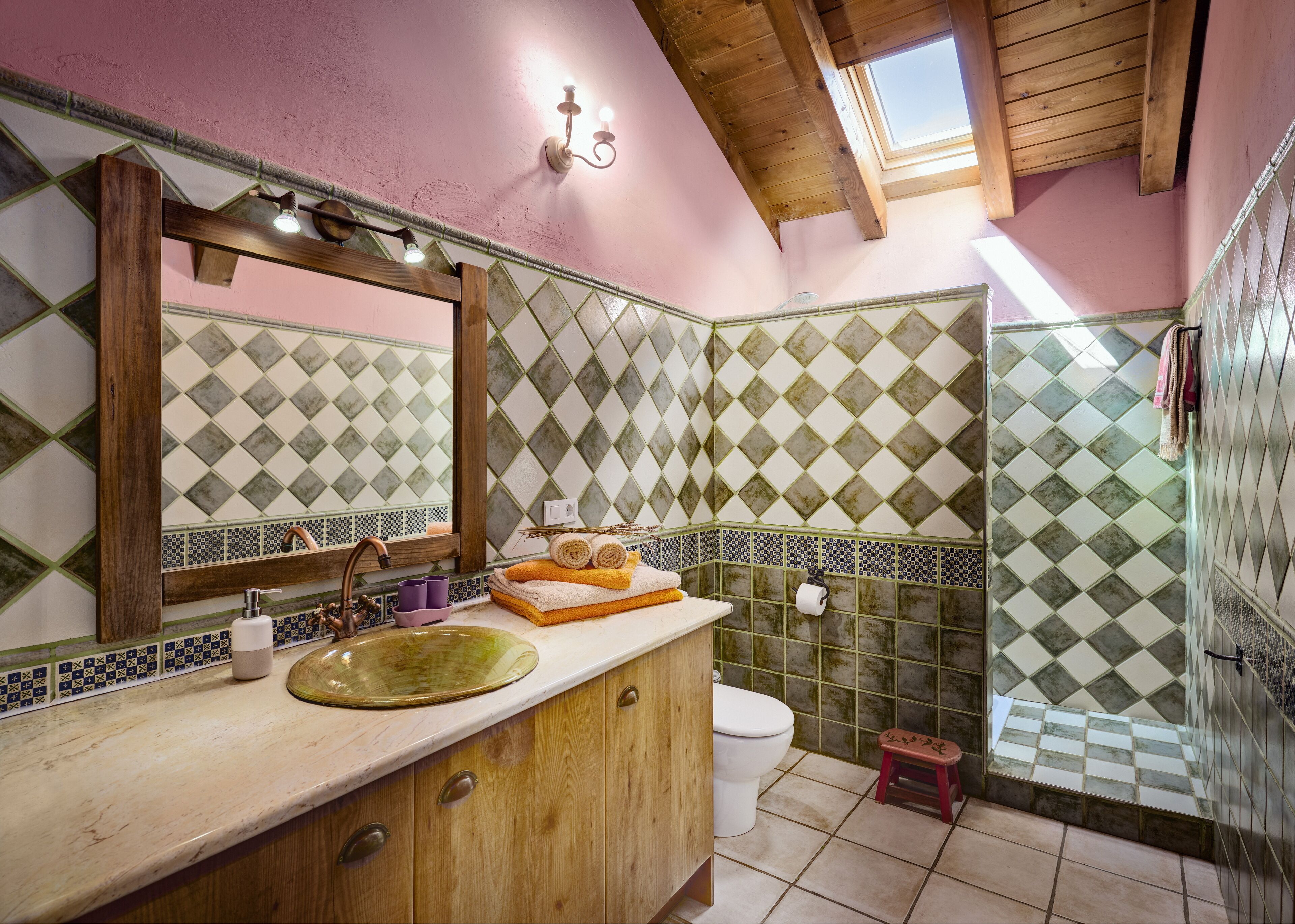Un baño con tocador de madera, un espejo grande, un lavamanos y un inodoro. Las paredes están cubiertas de azulejos con un diseño de diamante, y hay un tragaluz sobre el inodoro.