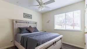 Une chambre à coucher comprenant un lit, un ventilateur de plafond, une fenêtre avec des stores et une œuvre d’art accrochée au mur.