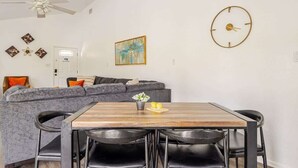 Een eetruimte met een houten tafel, zwarte stoelen, een grijze bank, een plafondventilator, een wandklok en een schilderij.