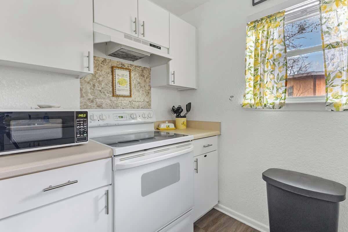 A compact kitchen with a microwave, stove, and sink. There is a trash bin and a window with curtains.