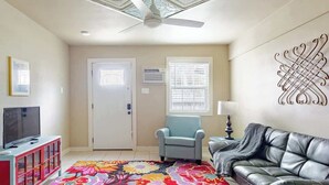 Un salon avec un ventilateur de plafond, un téléviseur posé sur un meuble, un canapé, un fauteuil et un tapis coloré.
