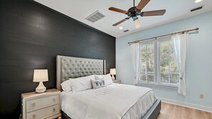 Une chambre à coucher avec une tête de lit capitonnée, un lit avec une literie blanche, une table de chevet avec une lampe, une fenêtre avec des stores et un ventilateur de plafond.