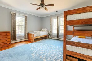A bedroom with a bunk bed, a wooden dresser, a bedside table with a lamp, and a large window with curtains.
