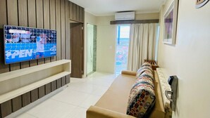 A living room with a flat-screen TV, a sofa, a window with curtains, and a door leading to another room. - Condomínio Piazza diRoma Acqua Park Splash (Caldas Novas)